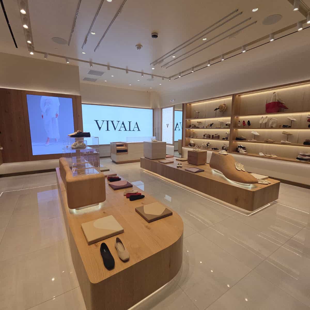 Modern retail store interior with wooden display tables, shelves with handbags and shoes, and a large screen displaying 'VIVAIA' branding.