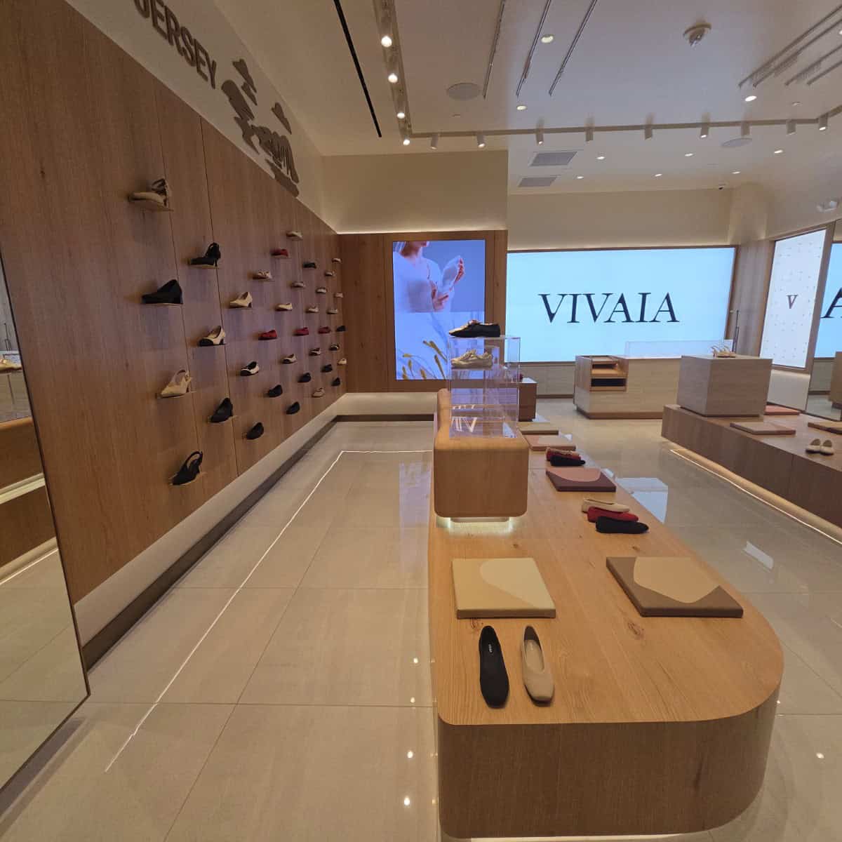 Shoe store interior with a wall display of various shoes, a central wooden table with shoe samples, and a large screen displaying 'VIVAIA' in the background.