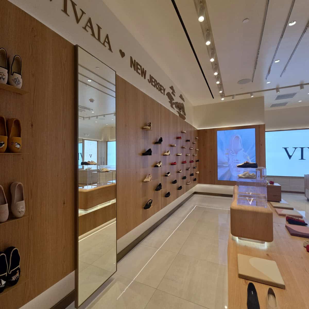 Interior of a shoe store with a wooden wall displaying various shoes, a large mirror, and a digital screen showing a model wearing shoes.