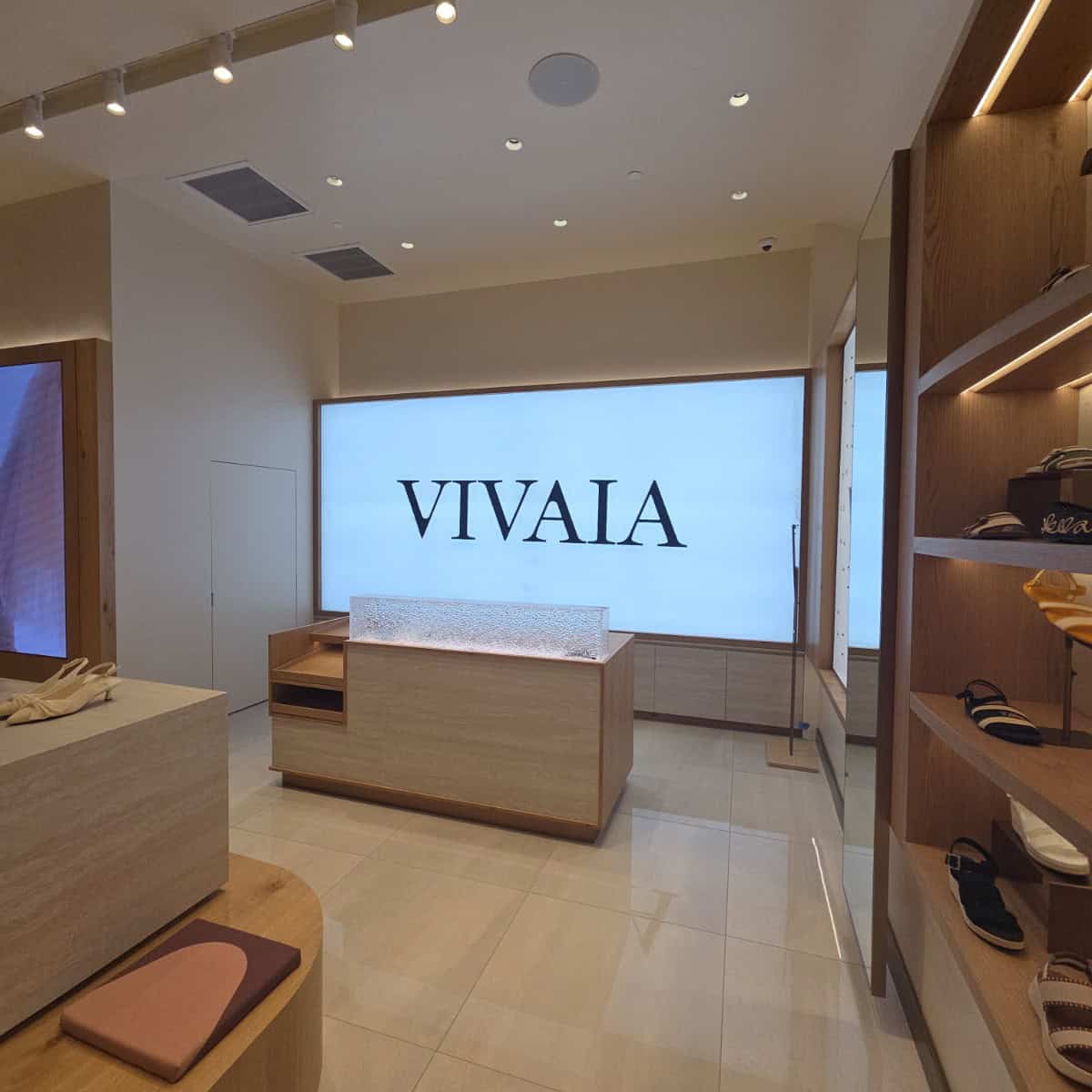 Interior of a retail store with a large illuminated sign reading 'VIVAIA' on the back wall, wooden shelves with shoes on the right, and a light-colored counter in the center.