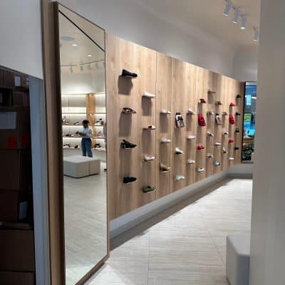 Display of various sneakers on a wooden wall in a shoe store with a mirror and customers in the background.